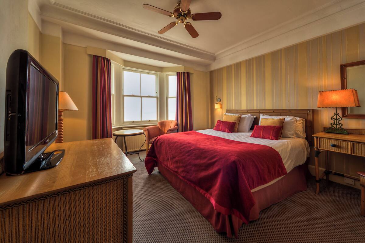 A hotel room with a large bed, a desk with a television, a ceiling fan, and a window with curtains.