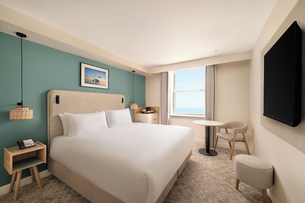 A hotel room with a large bed, bedside tables, a small round table, a chair, a TV mounted on the wall, and a window with curtains.