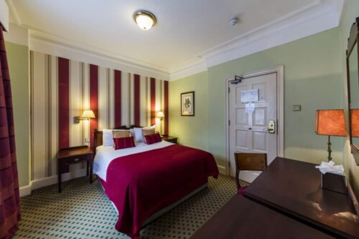 A hotel room with a large bed, a desk, a chair, a nightstand, and a door.