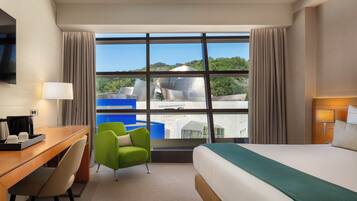 Una habitación de hotel con un ventanal grande, una cama, un escritorio, una silla y un televisor.