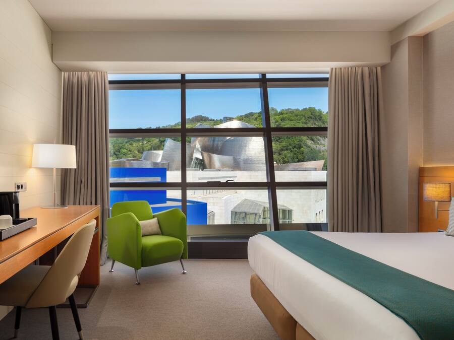 Una habitación de hotel con un ventanal grande, una cama, un escritorio, una silla y un televisor.
