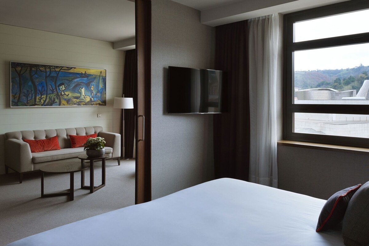 A hotel room with a bed, a sofa, a TV, a painting, and a window with a view.