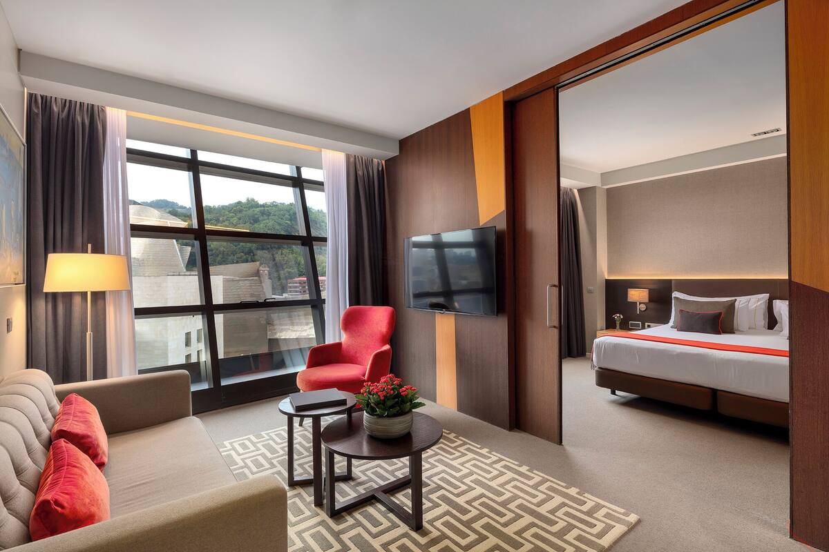 Un hotel moderno con sofá, sillón rojo, una pequeña mesa con un jarrón de flores, cama con detalles en rojo, televisor de pantalla plana montado en la pared y un amplio ventanal con vista a la ciudad.