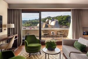 Una moderna camera d'albergo con divano, sedie, un tavolino con una pianta e un balcone che offre vista su un edificio e sul verde.