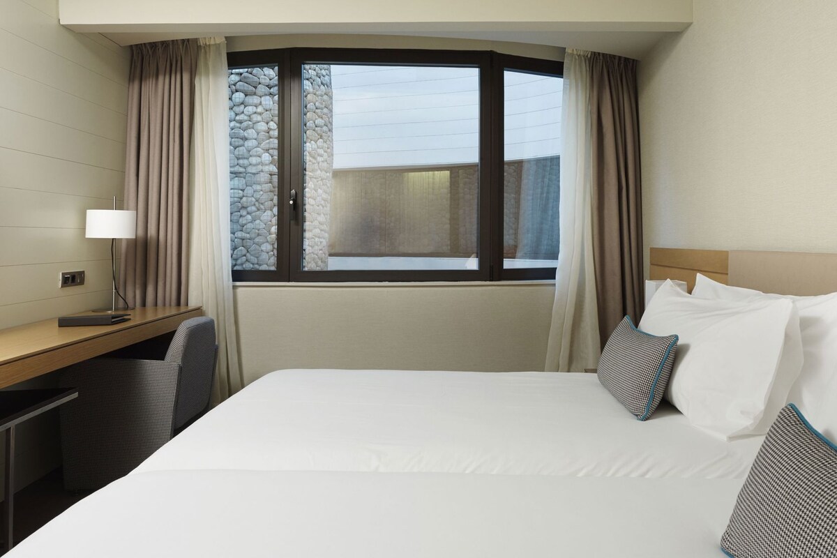 A hotel room with a bed, a desk, a chair, a lamp, and a window with curtains.