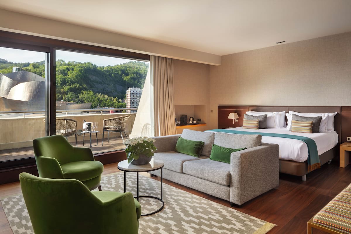 Habitación de hotel con una cama grande, un sofá, dos sillones, una mesita con una planta y vistas a la ciudad y las colinas.