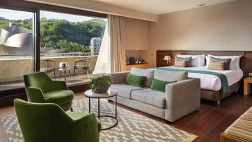 Habitación de hotel con una cama grande, un sofá, dos sillones, una mesita con una planta y vistas a la ciudad y las colinas.