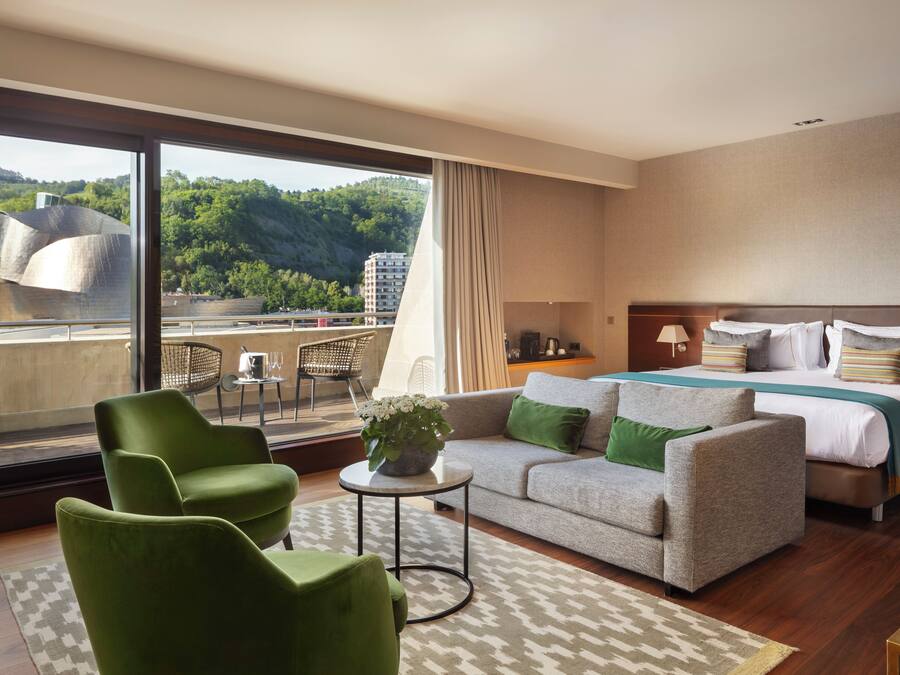 Habitación de hotel con una cama grande, un sofá, dos sillones, una mesita con una planta y vistas a la ciudad y las colinas.