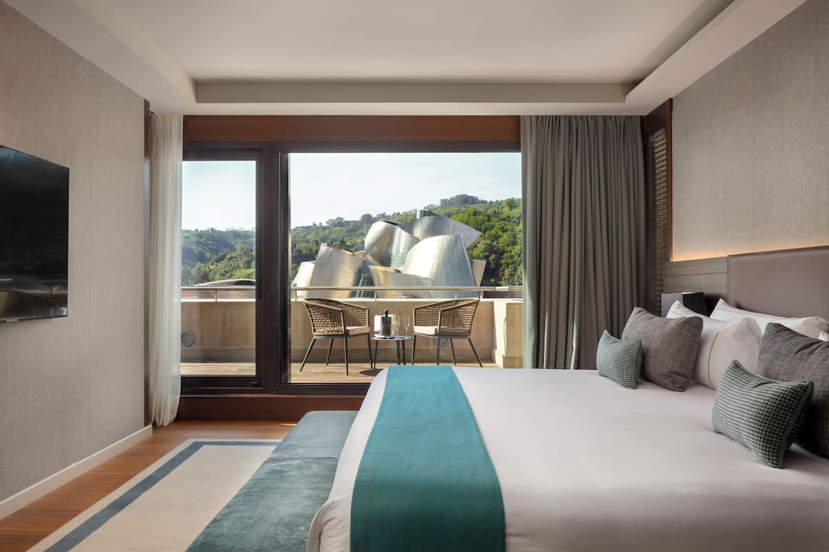 Habitación de hotel con una cama grande, televisión, balcón con mesa y sillas, y vista al exterior.
