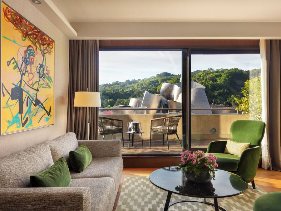 Quarto de hotel moderno com varanda, sofá, poltrona verde, mesa com um vaso de flores e uma grande pintura abstrata na parede.