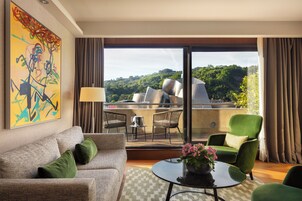 Una moderna camera d'albergo con balcone, un divano, una poltrona verde, un tavolo con un vaso di fiori e un grande quadro astratto appeso alla parete.