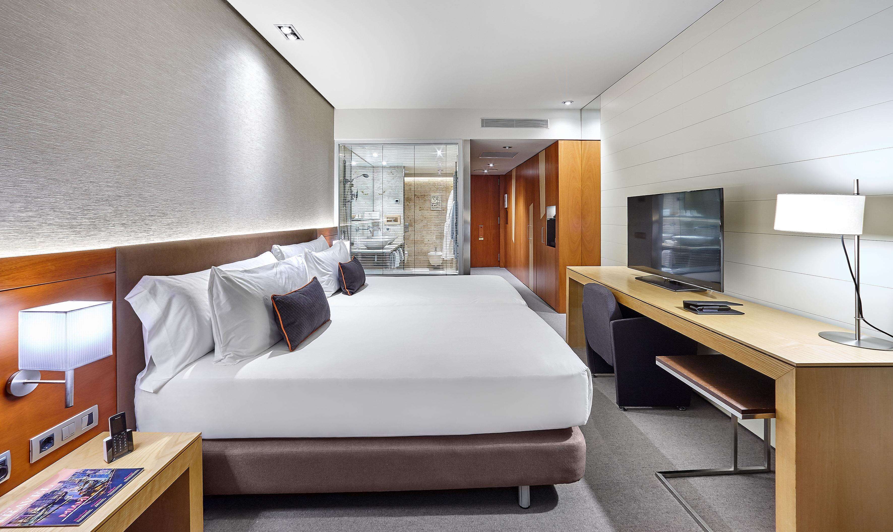 Habitación de hotel moderna con una cama grande, un escritorio, un televisor y un baño visible a través de una mampara de vidrio.