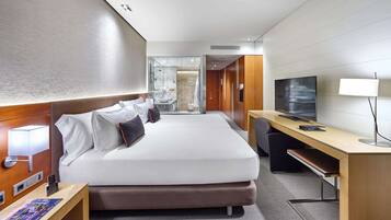 Habitación de hotel moderna con una cama grande, un escritorio, un televisor y un baño visible a través de una mampara de vidrio.