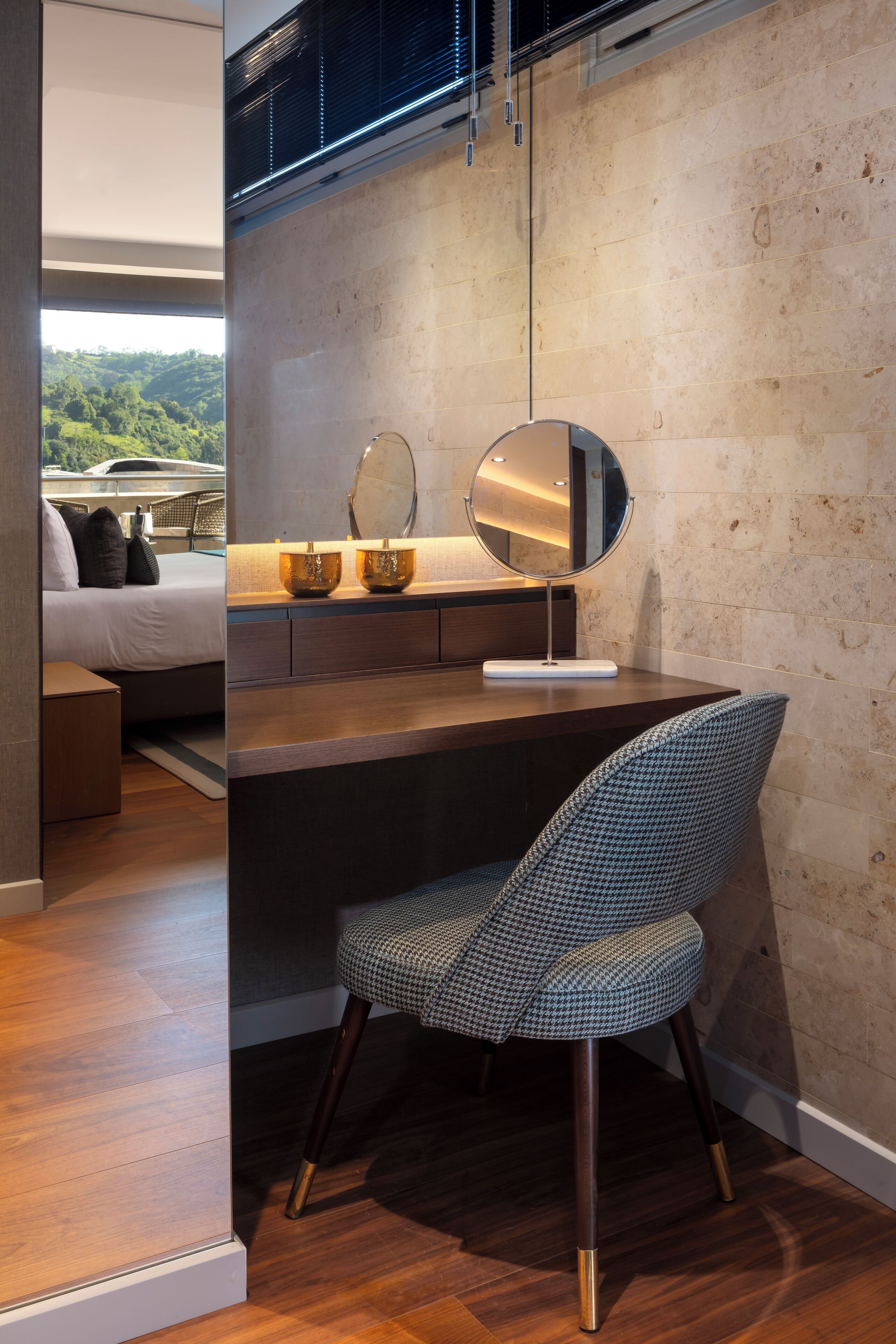 Un dormitorio moderno con un escritorio de madera, una silla estampada de piedritas, un espejo redondo y una cama con vistas al exterior.