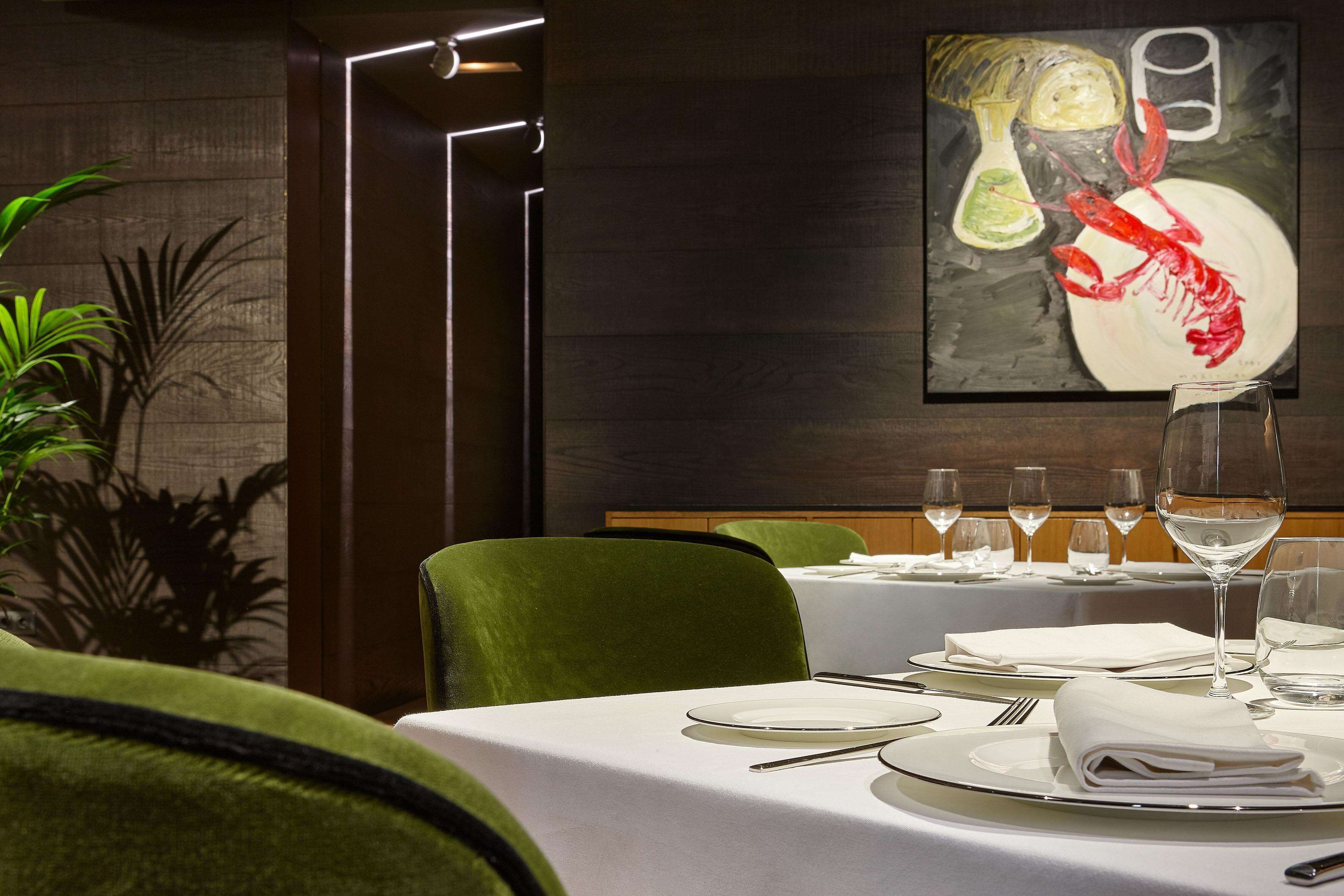 A dining area with a table set for two, a painting on the wall, and a plant in the corner.