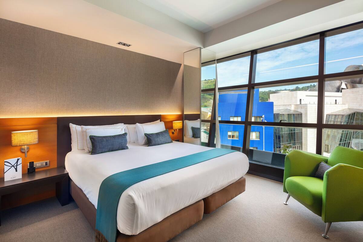 Una habitación de hotel moderna con una cama grande, un sillón verde y vistas a edificios a través de amplios ventanales.