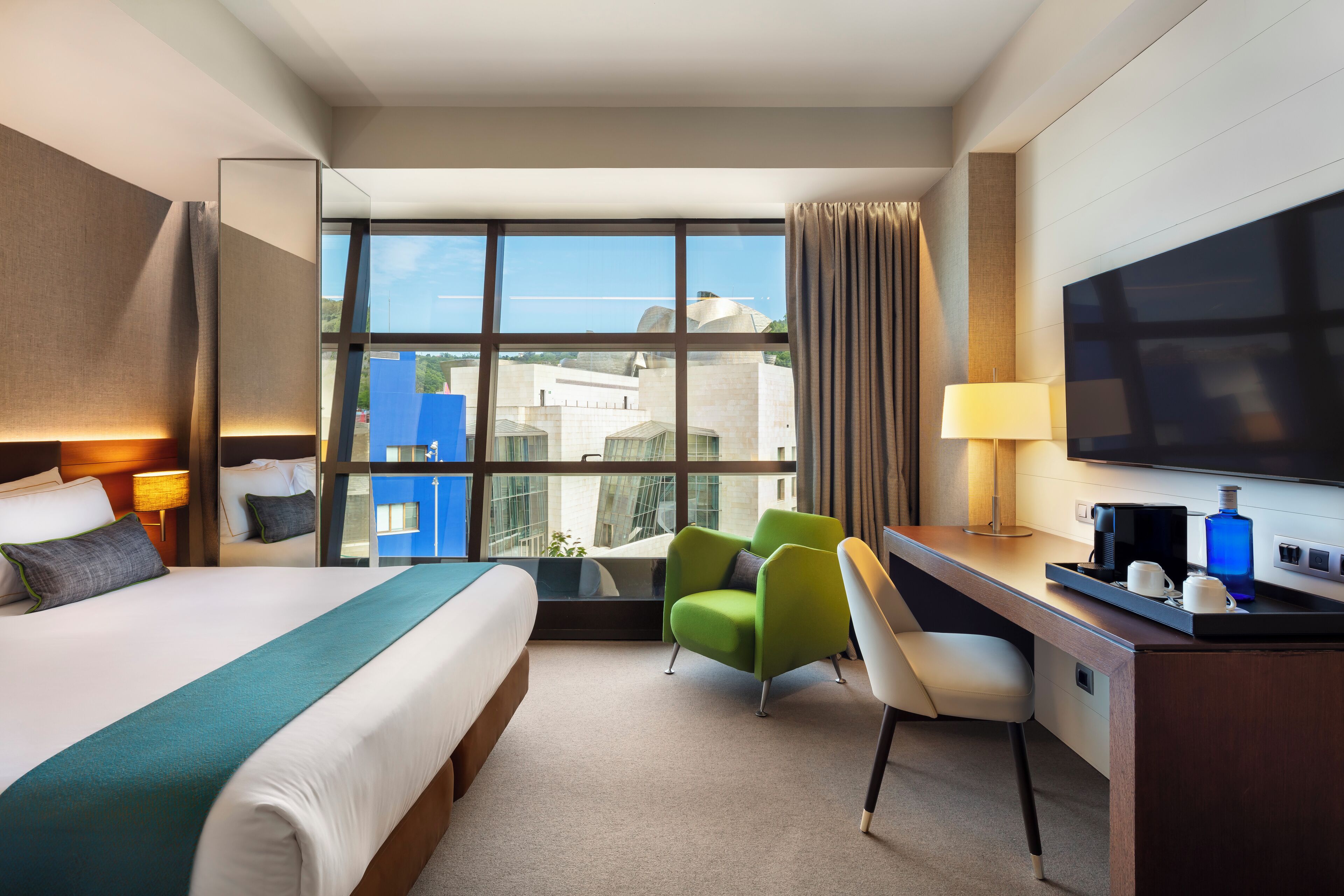 A hotel room with a large bed, a desk with a chair, a television, and a view of buildings through large windows.