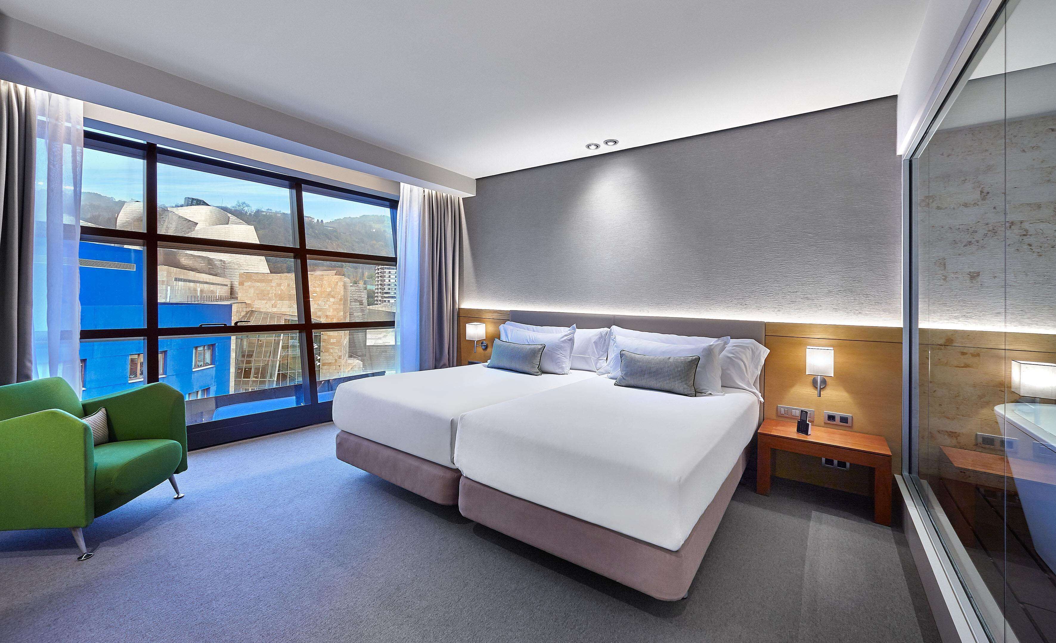 Habitación de hotel con una cama grande, un sillón verde, una mesita de noche y vistas a edificios a través del ventanal.