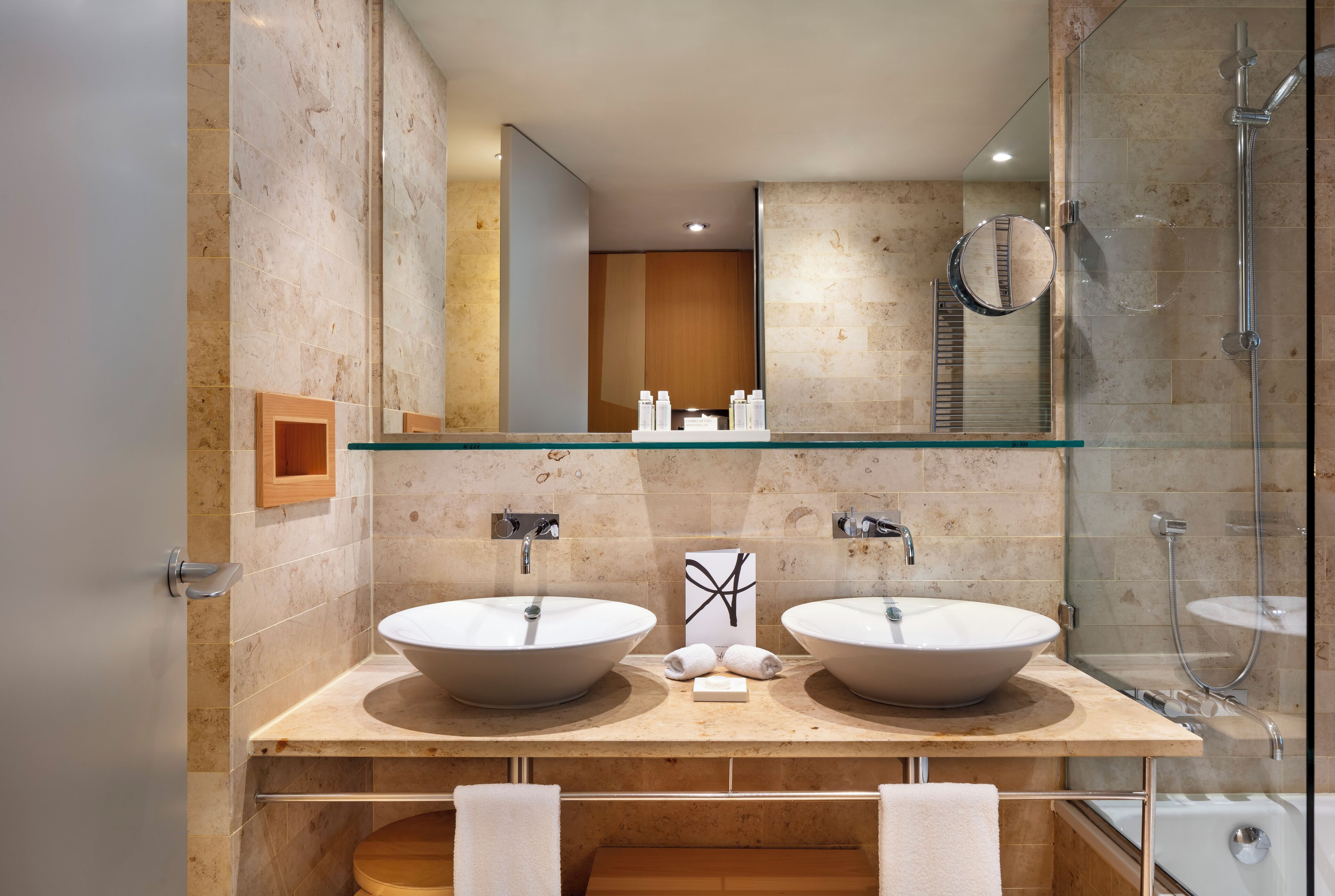 Un baño moderno con dos lavamanos grandes, cabina de ducha con vidrio y tocador de madera.