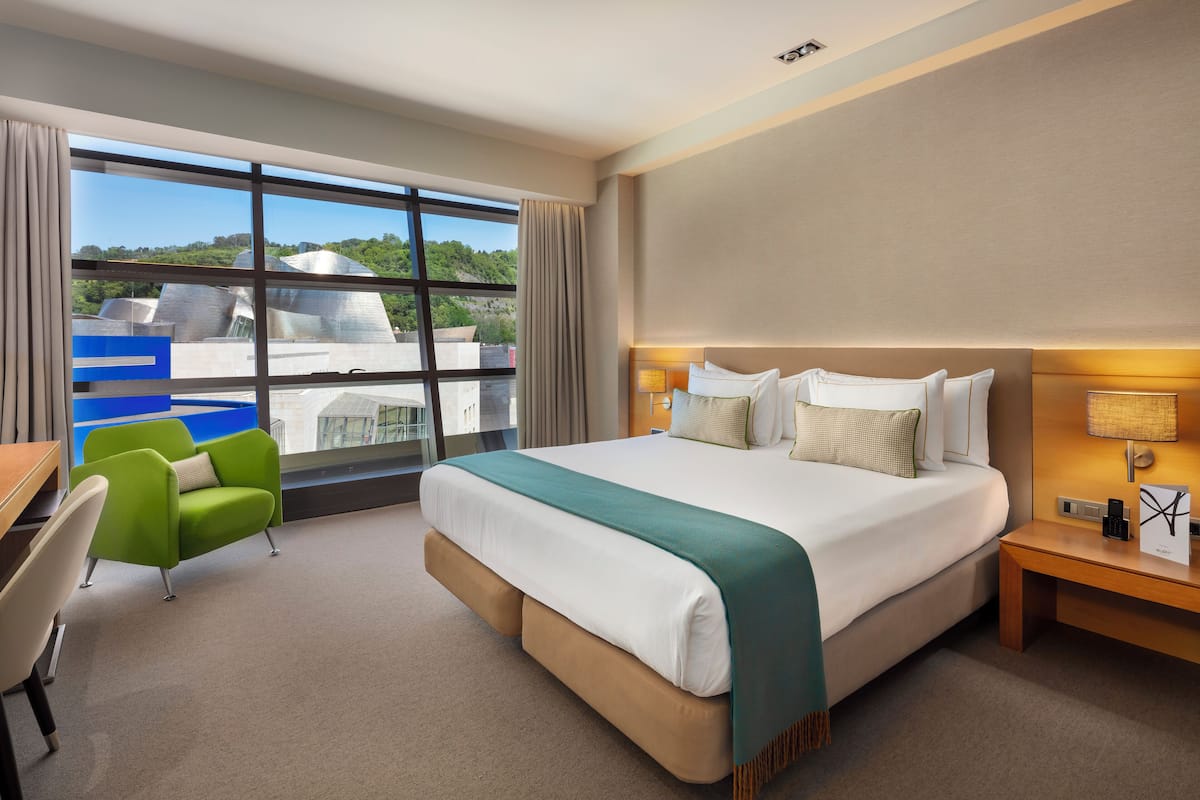 Habitación de hotel con una cama grande, un escritorio, una silla verde y vistas a un edificio a través de amplios ventanales.