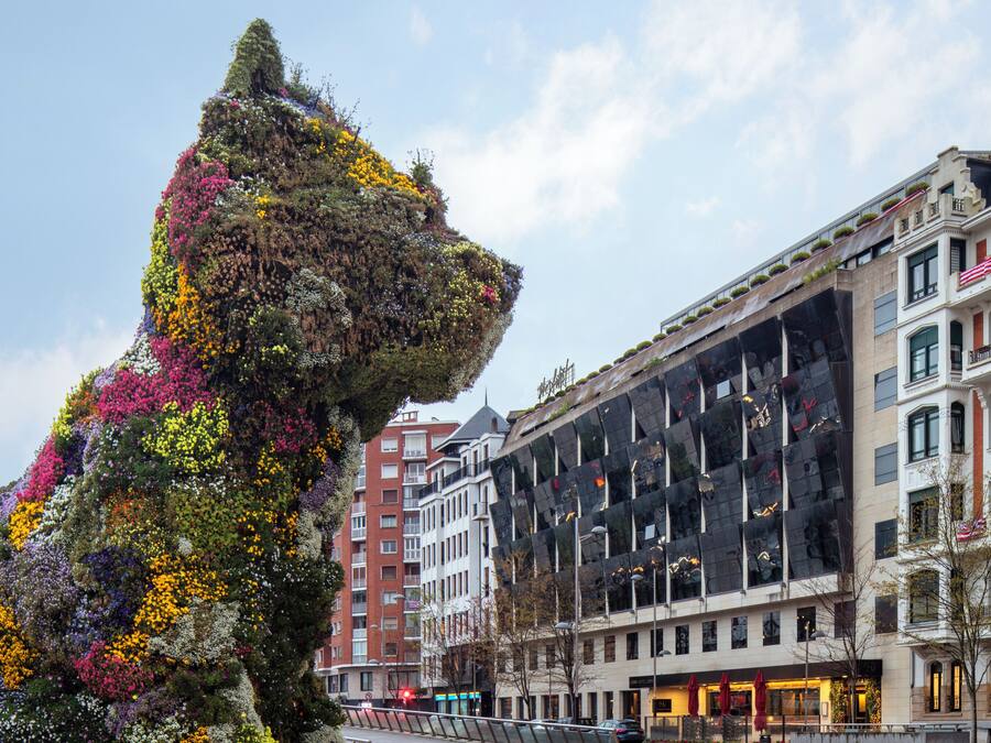 Una gran escultura floral con forma de cabeza de león en una plaza urbana con edificios modernos al fondo.
