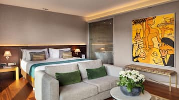 Habitación de hotel con cama, sofá, una pequeña mesa con una planta y un cuadro abstracto grande en la pared.