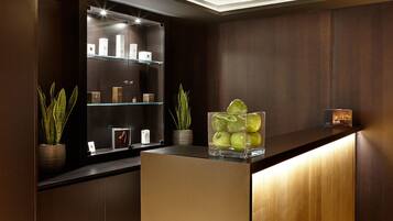 Un área de recepción con mostrador de madera, estanterías de cristal, plantas en macetas y un recipiente de vidrio con manzanas.