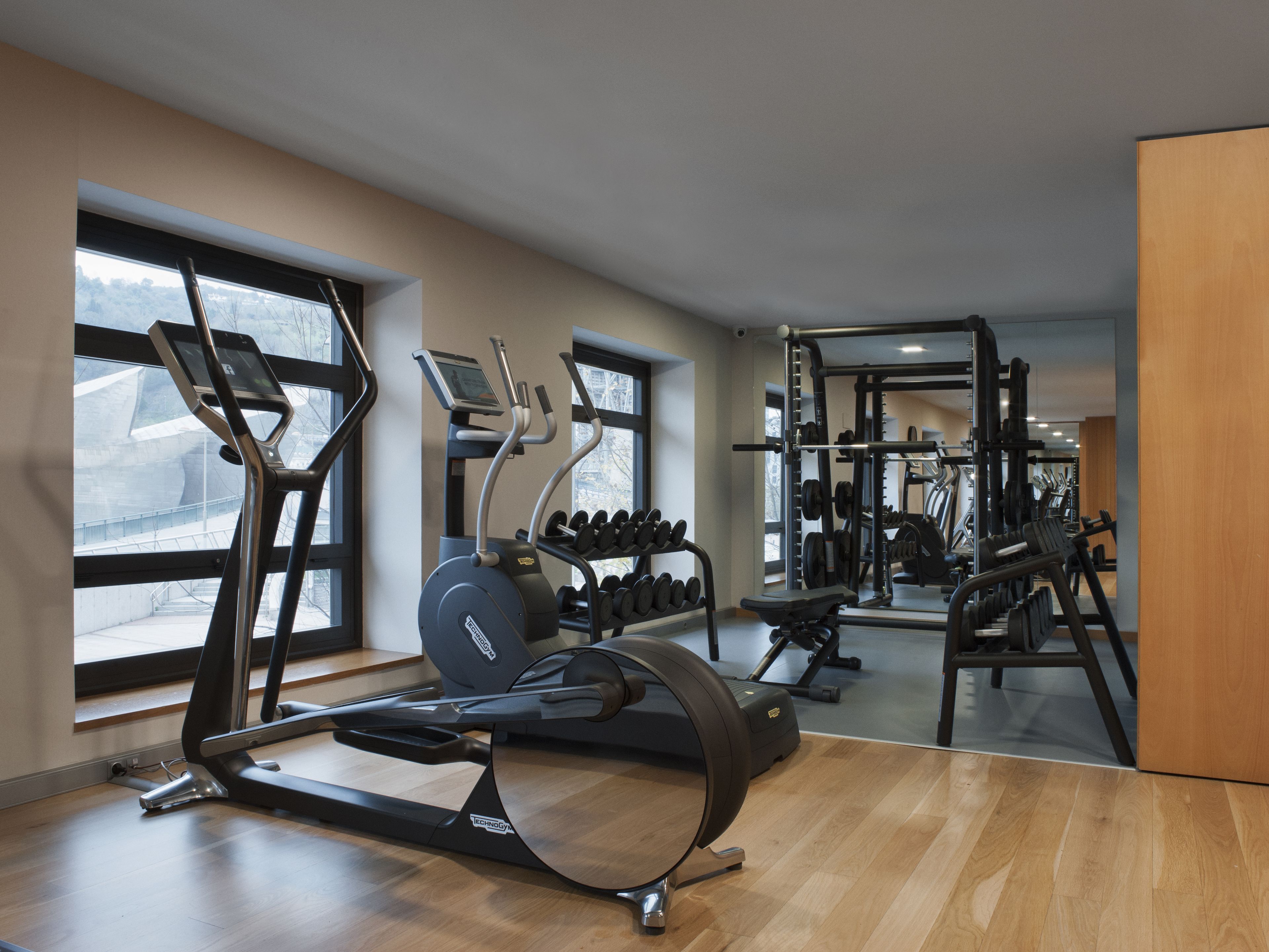 A modern gym with cardio machines, weightlifting equipment, and large windows offering views of the outdoors.
