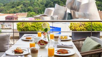 Mesa de desayuno con diferentes platos, bebidas y una rosa en un jarrón, con vistas a un paisaje urbano y un edificio moderno.