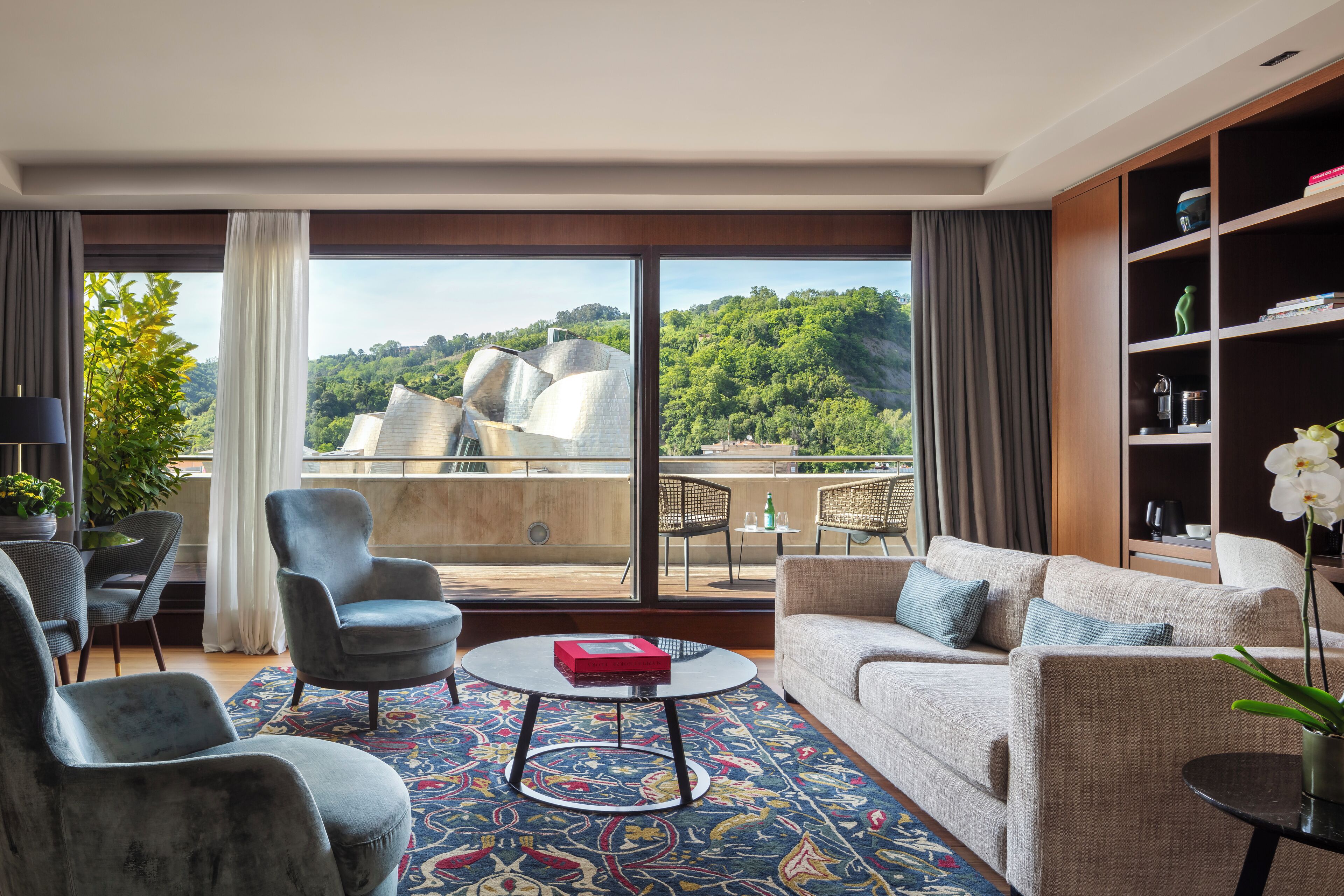 Una sala de estar moderna con un ventanal grande, un sofá, sillones, una mesa de centro y una estantería.