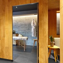 Una stanza spa con pavimento in legno, un lettino per massaggi, un accappatoio bianco appeso e una pianta in vaso.