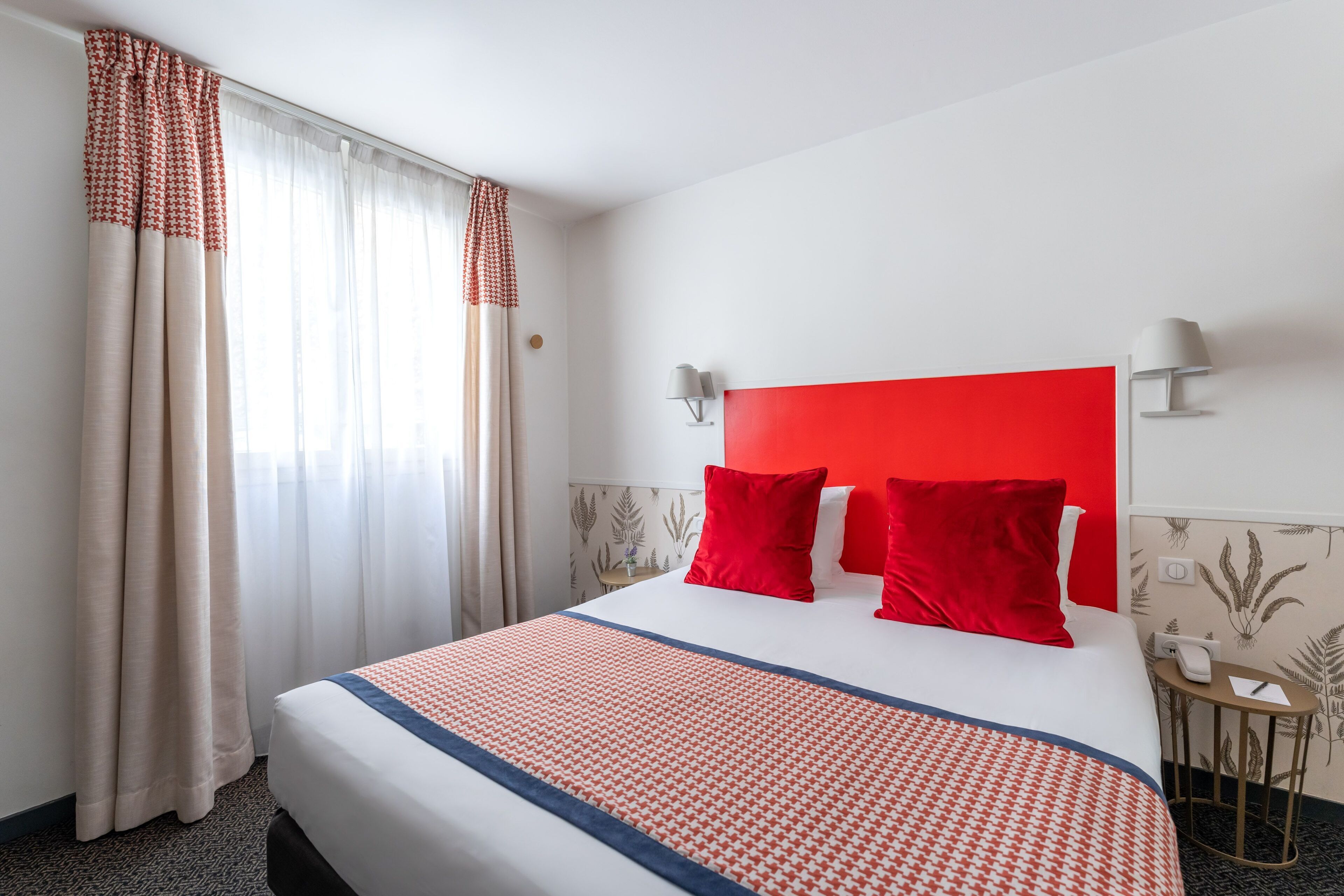 A hotel room with a large bed, red and white bedding, a red headboard, two bedside lamps, a small table with a phone, and a window with curtains.