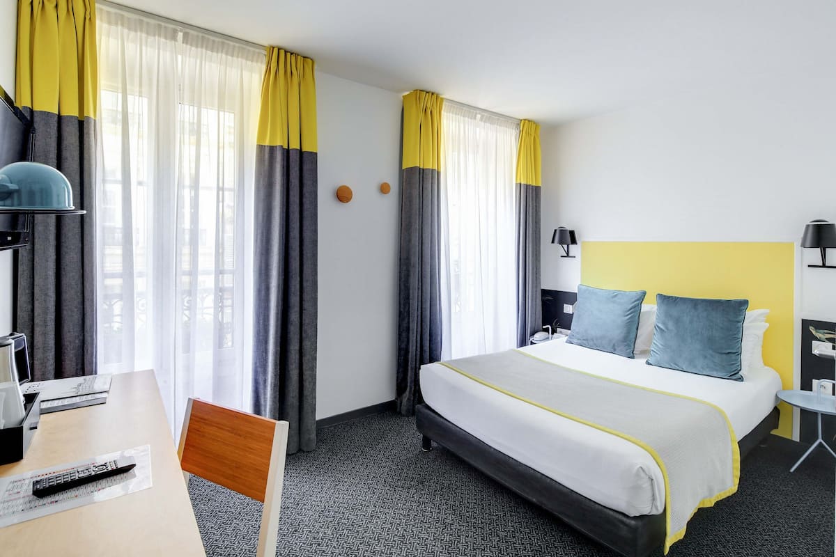 Una habitación de hotel con cama, escritorio, silla, lámpara y cortinas.
