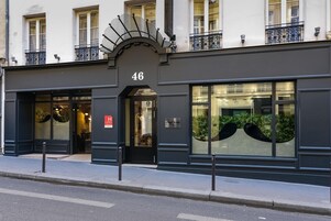 Un immeuble à la façade sombre, portant le numéro 46, avec de grandes fenêtres ornées de plantes, une entrée bien visible et un petit balcon doté d’une balustrade en fer forgé.
