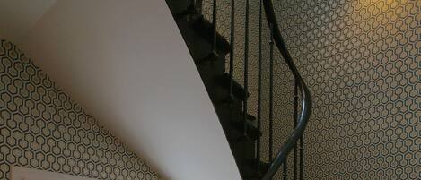 Una escalera de madera con barandilla negra, un ventanal con vista a edificios y papel tapiz estampado.
