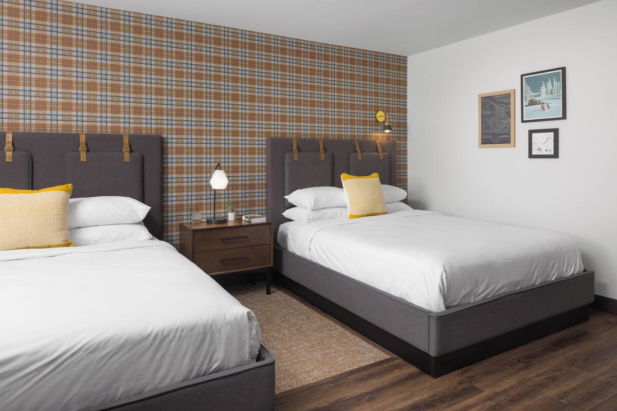 Habitación de hotel con dos camas, una mesita de noche, una lámpara y una pared con estampado de cuadros.