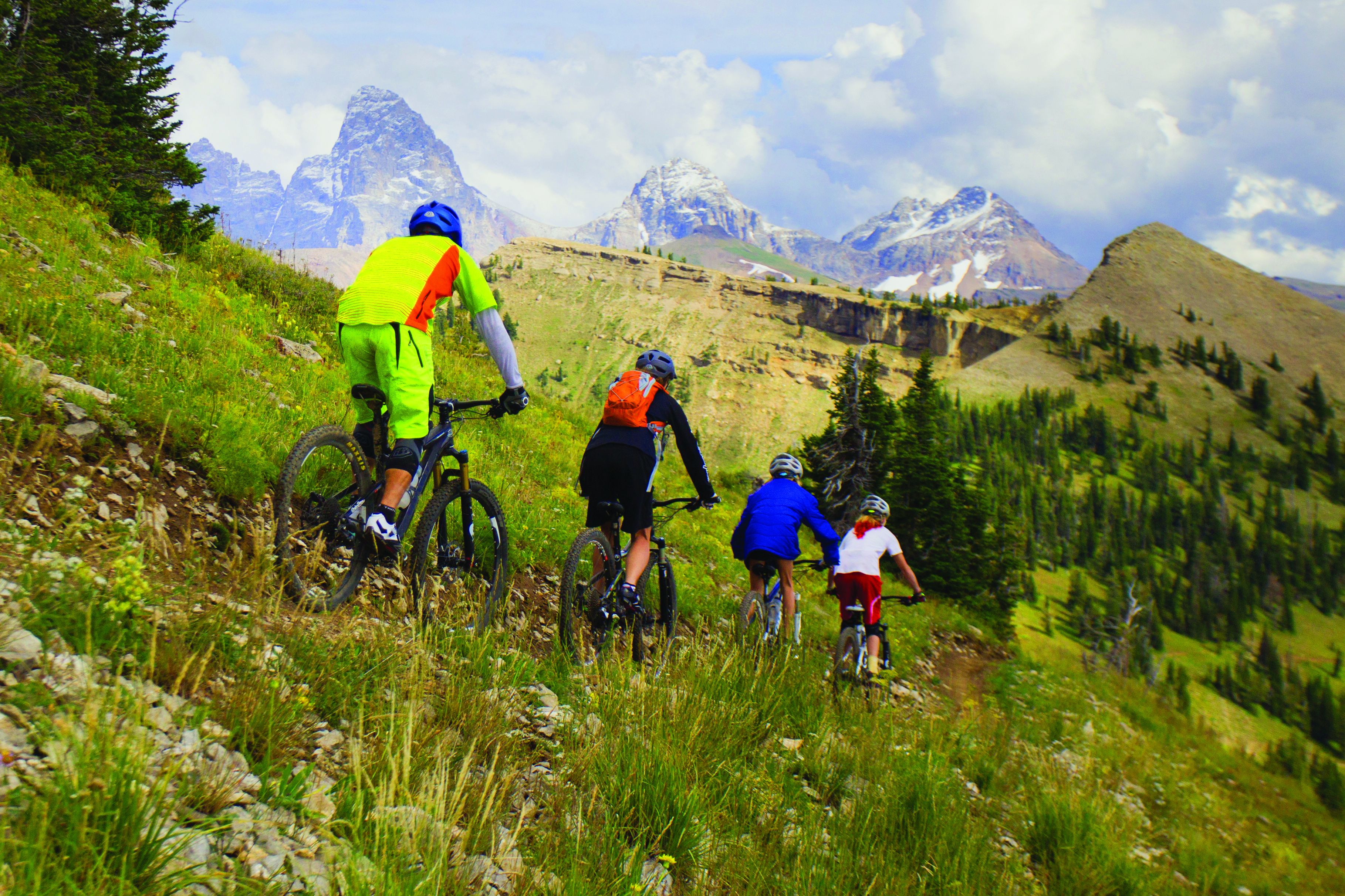 A group of people on mountain bikes on a grassy trail with mountains in the background.