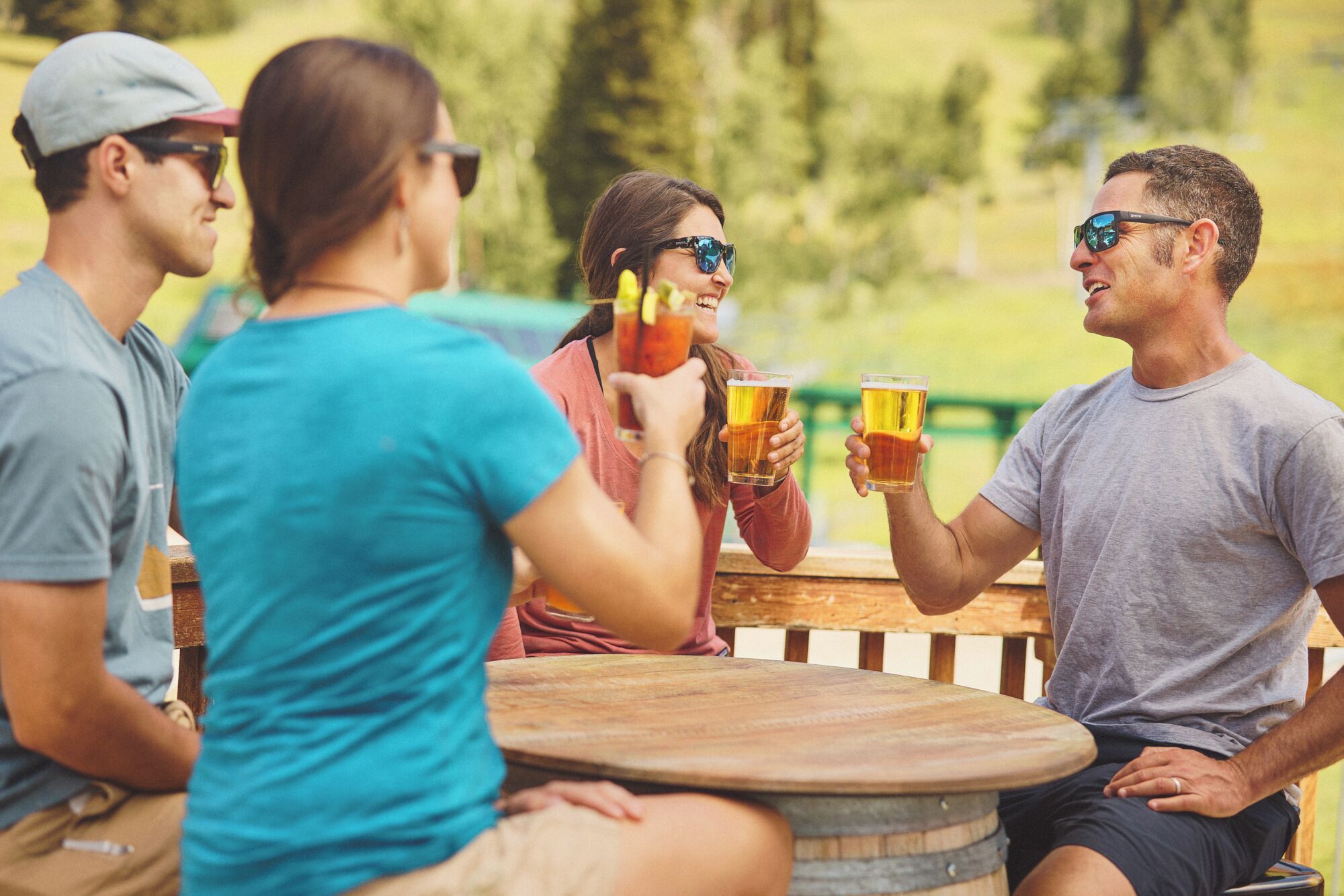A group of people sitting around a wooden table outdoors, holding glasses of beer.
