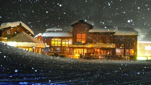 Un bâtiment recouvert de neige, avec une entrée illuminée et un espace extérieur aménagé avec des si èges, le tout entouré de neige.