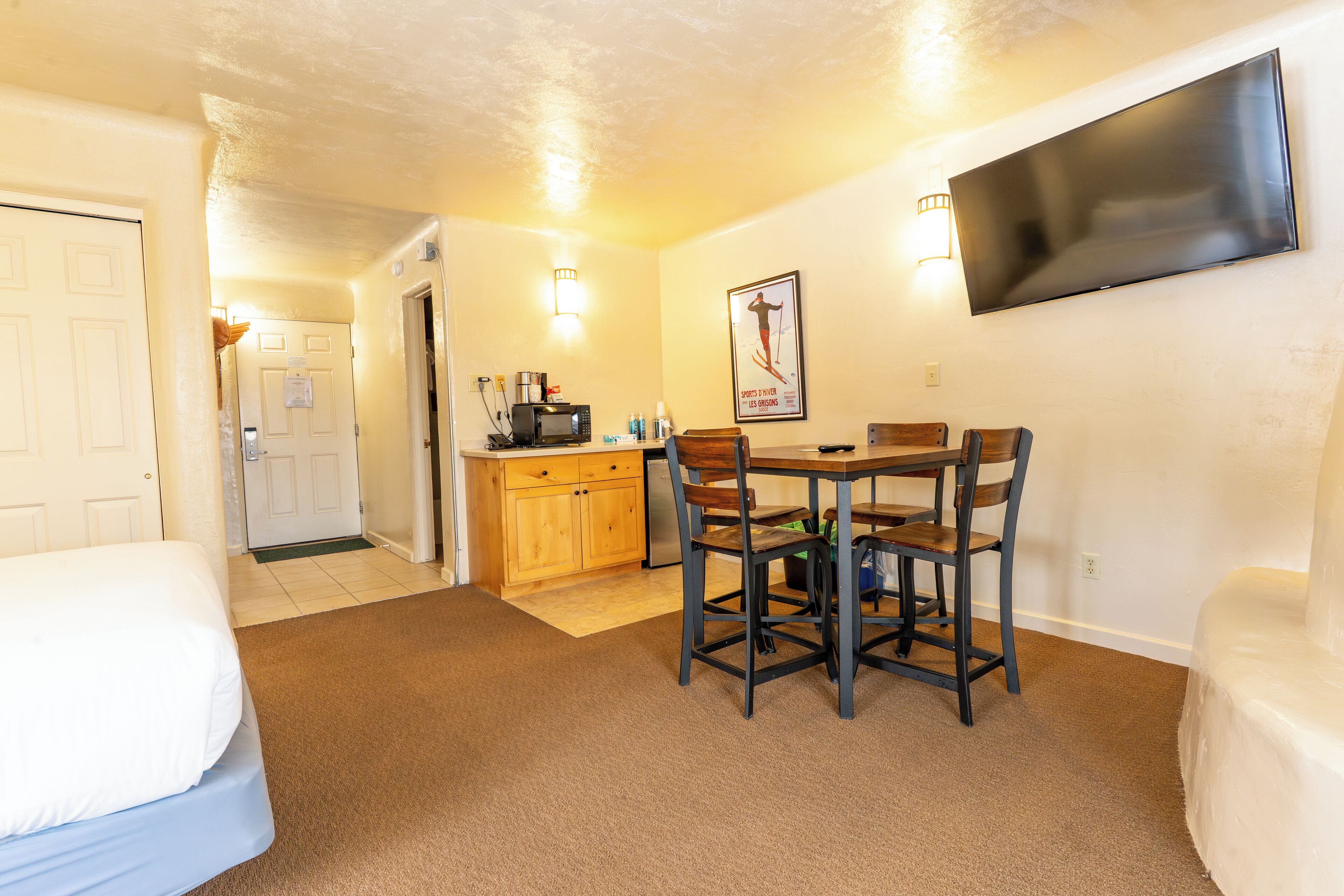A hotel room with a bed, a dining table with chairs, a kitchenette, and a TV mounted on the wall.