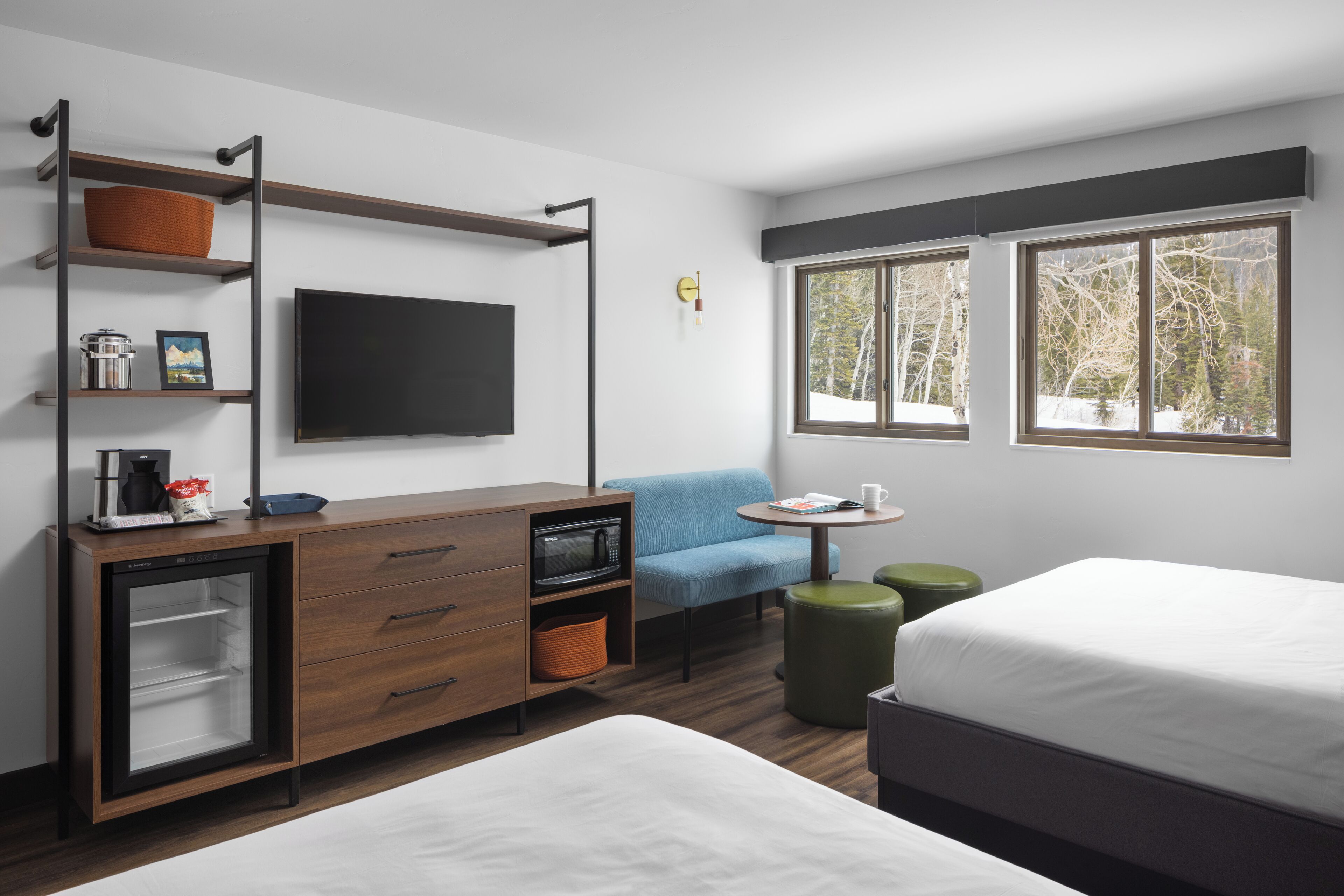 A modern hotel room with a bed, a desk with a flat-screen TV, a microwave, and a small refrigerator. There are two windows with a view of trees and a chair with a table.