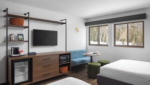 A modern hotel room with a bed, a desk with a flat-screen TV, a microwave, and a small refrigerator. There are two windows with a view of trees and a chair with a table. - Grand Targhee Resort (Alta)