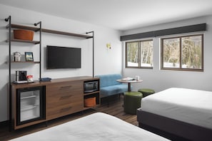 A modern hotel room with a bed, a desk with a flat-screen TV, a microwave, and a small refrigerator. There are two windows with a view of trees and a chair with a table.