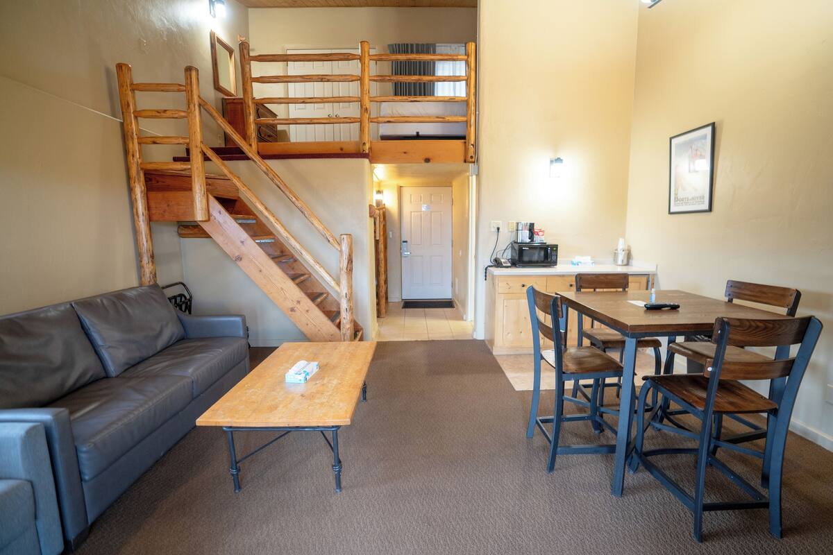 Una habitación con una escalera de madera, un sofá, una mesa y una zona de comedor con sillas.