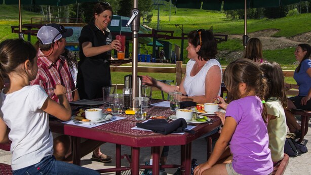 Un groupe de personnes est assis à une table en plein air, dont le cadre est en métal et qui est surmontée d’une sorte de parasol vert.