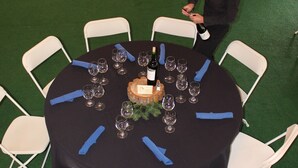 Une table ronde, entourée de chaises, avec une bouteille de vin et des verres, le tout posé sur un sol recouvert d’une moquette verte.