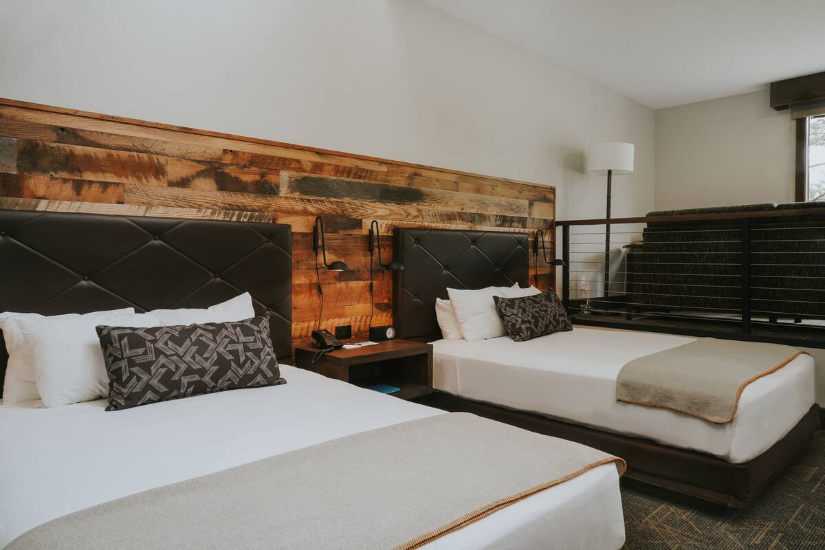 A hotel room with two beds, a wooden headboard, a bedside table with a lamp, and a window with a view.
