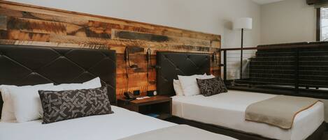 Habitación de hotel con dos camas, cabecero de madera, mesita de noche con lámpara y ventana con vistas.