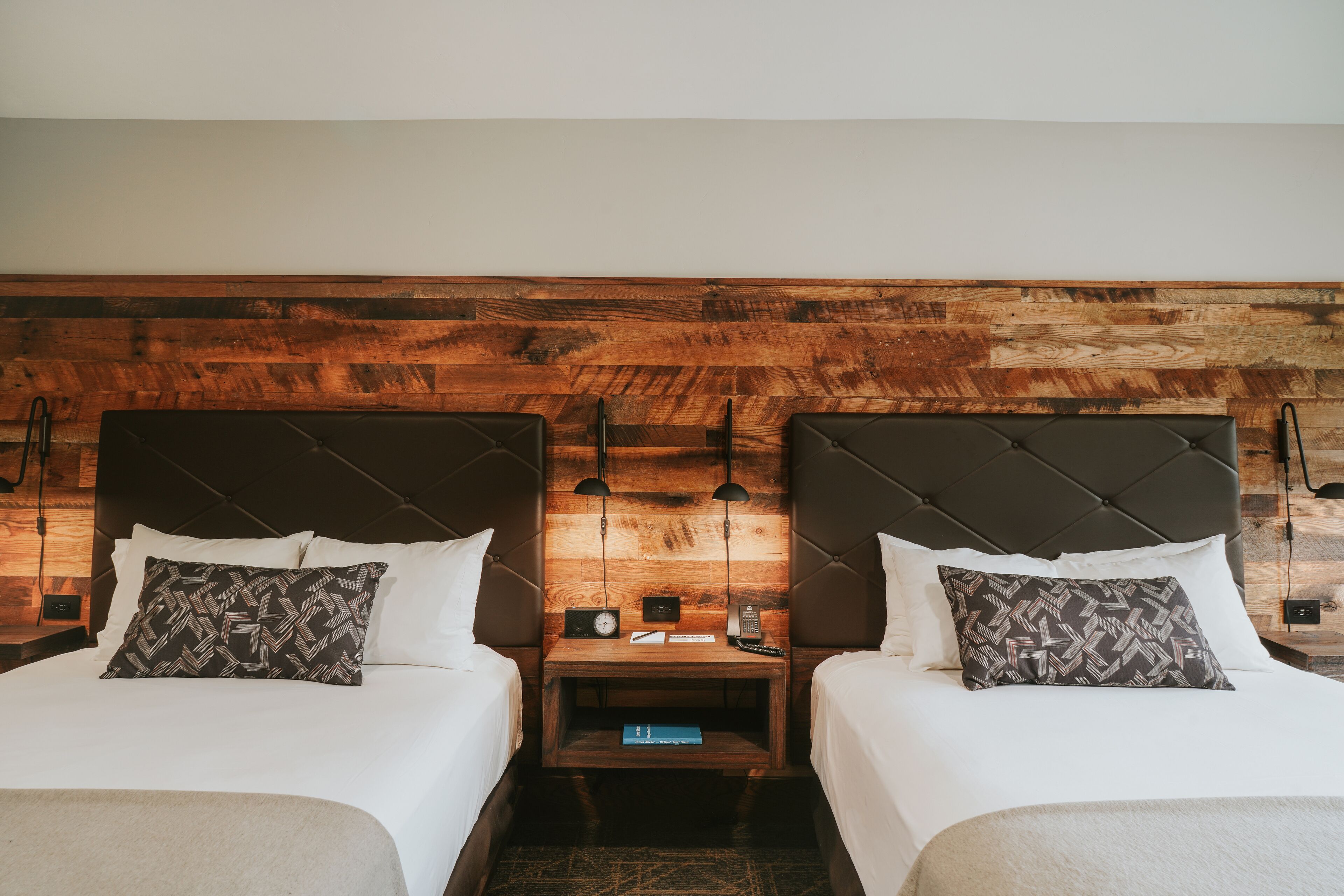 A hotel room with two beds, a wooden headboard, bedside tables with lamps, and a small shelf with books.
