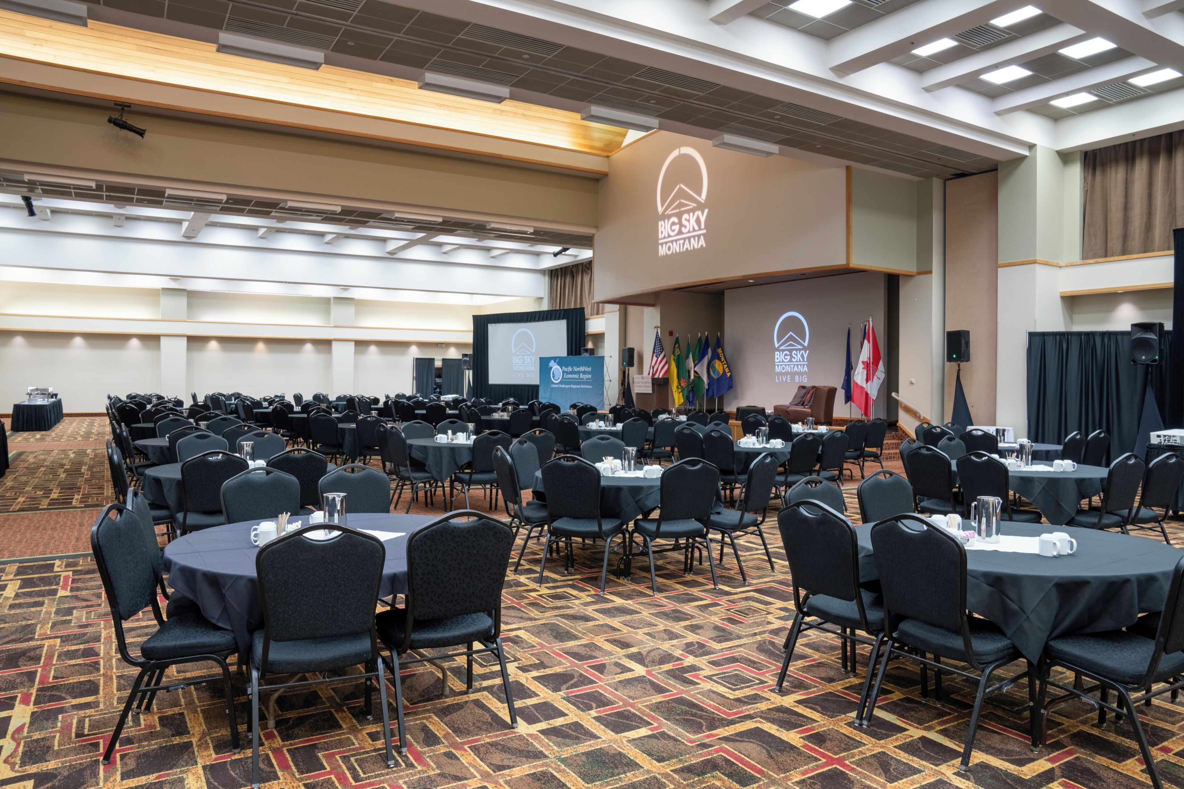 Une salle de conférence avec des tables rondes, des chaises noires et une scène équipée d’un écran de présentation. Des drapeaux sont accrochés au mur.